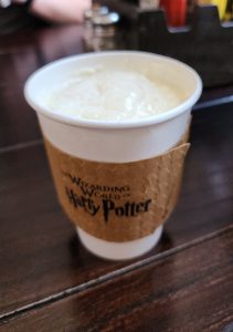 A frothy cup of hot creamy butterbeer in a white cup wrapped in a brown cardboard protective sleeve.