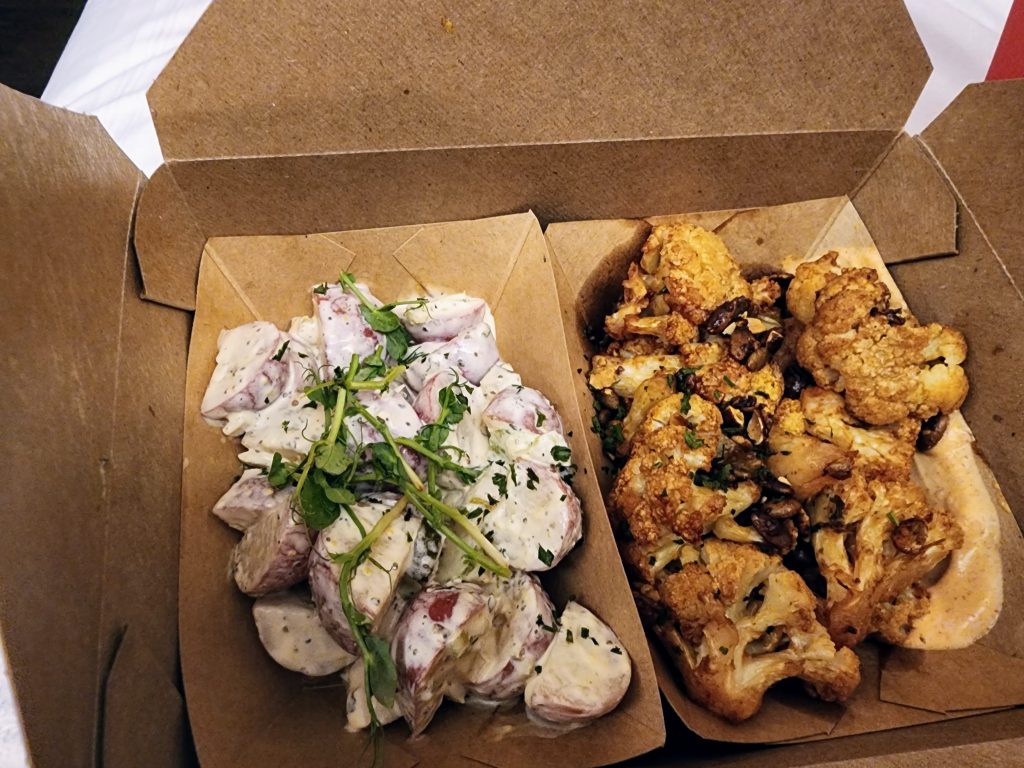 A container of two sides: potato salad and a barbecue cauliflower with sauce and pepitas.