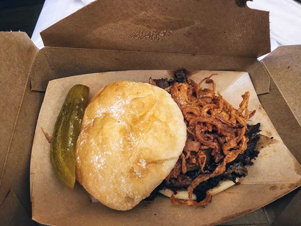 A sandwich filled with barbecue brisket, onion strings and a pickle on the side.