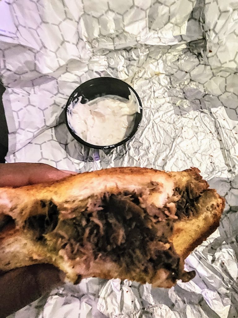 A half-eaten brisket sandwich with a small container of white sauce.