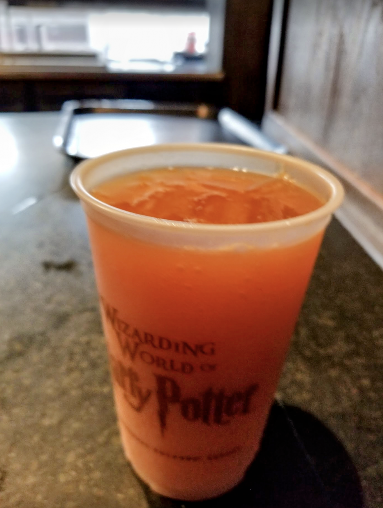 A cup of iced brown-orange colored pumpkin juice in a clear Harry Potter themed cup.